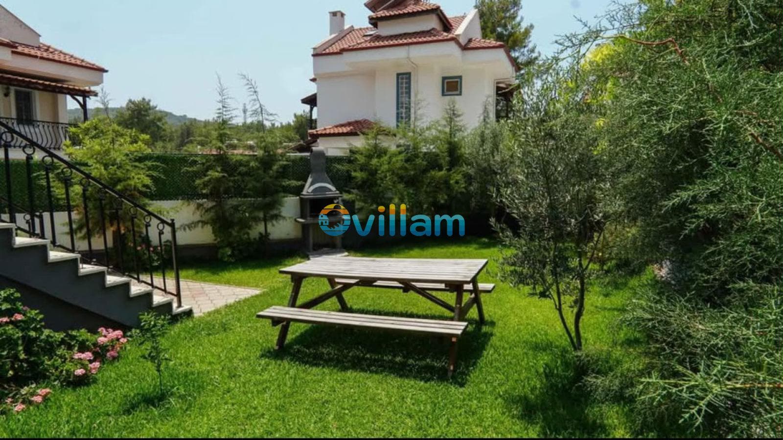 Bahçede ahşap piknik masası, taş barbekü ve yeşil bitkiler yer alıyor.