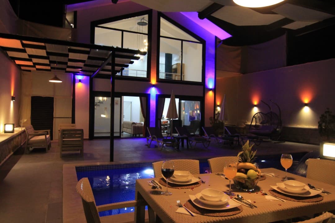 Gece ışıklandırmalı havuz kenarında masa, havuz ve şezlongların görüldüğü villa cephesi.