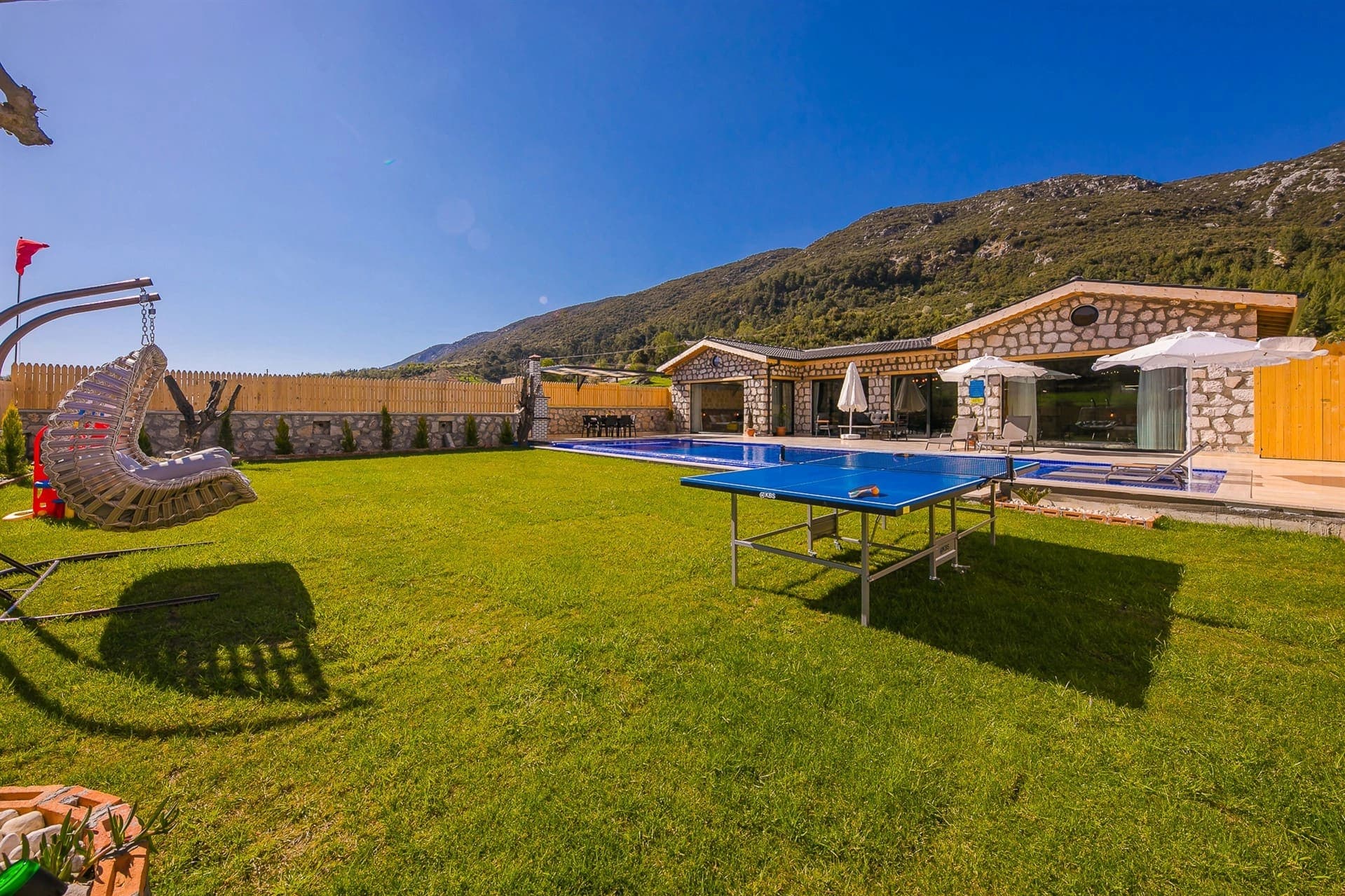Çim bahçesinde havuz, masa tenisi ve salıncak bulunan taş villa.