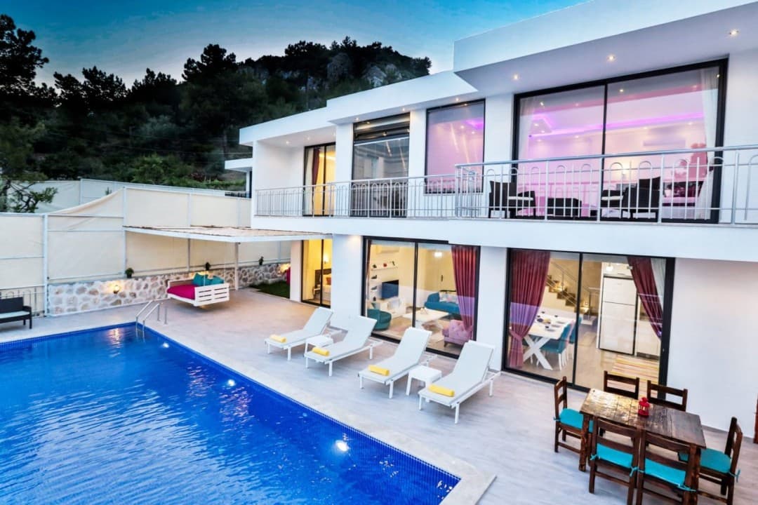 Havuz başında şezlonglar ve yemek masası bulunan iki katlı, aydınlatılmış modern beyaz villa.