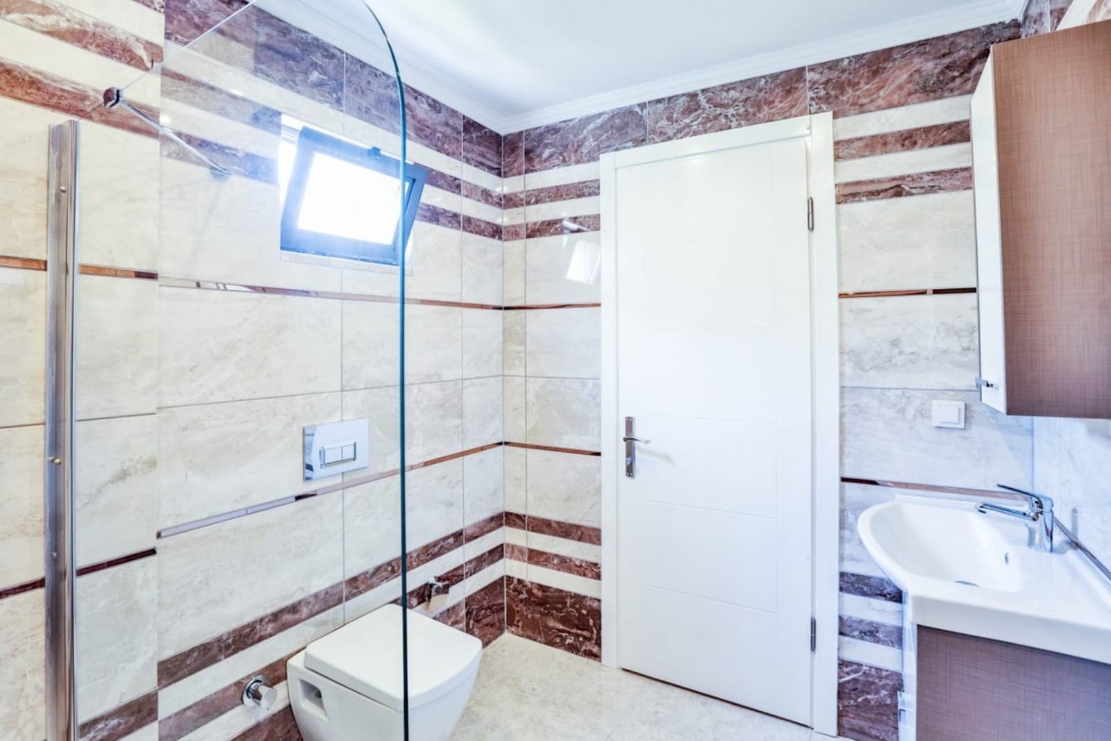 Kahverengi mermer detaylı duvarlara sahip, duşakabinli ve beyaz kapılı banyo.