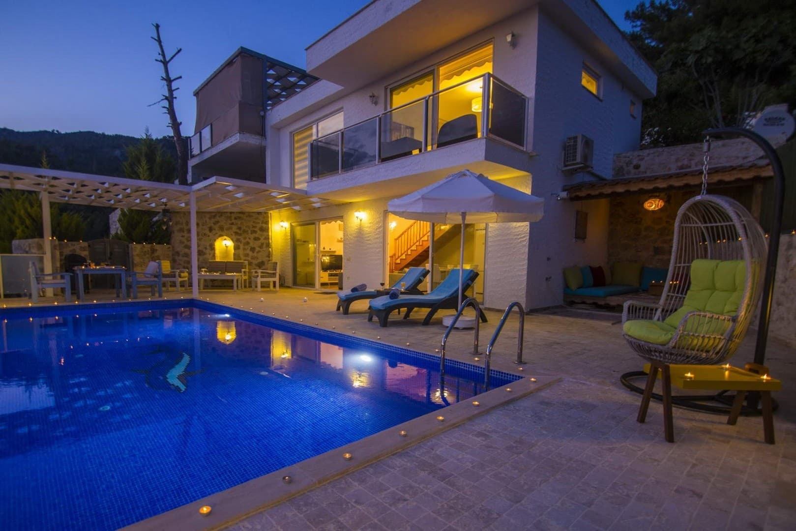 Akşam vakti aydınlatılmış, havuz başında şezlonglar, salıncak ve oturma grubu bulunan iki katlı villa.