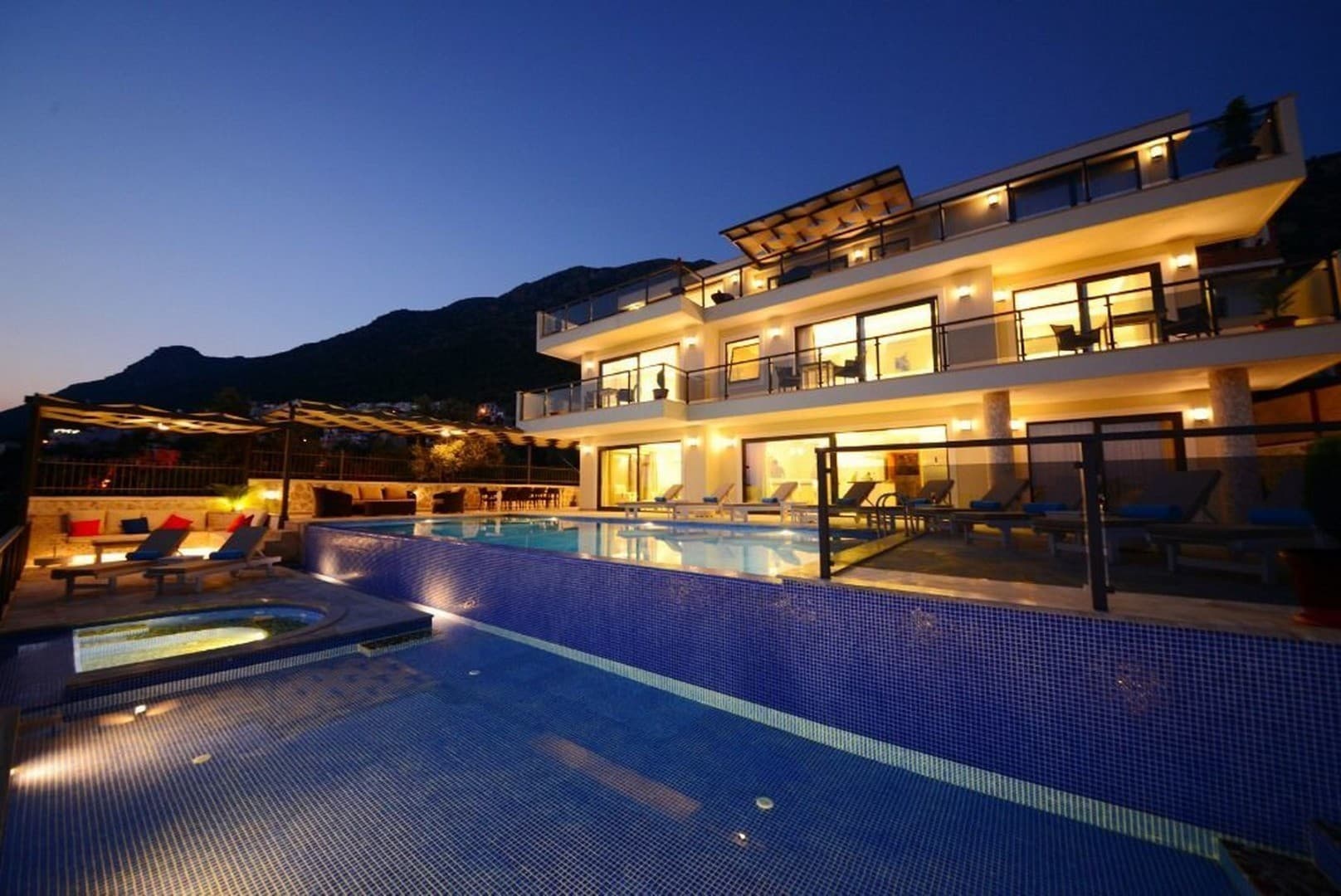 Akşam vaktinde ışıklandırılmış, sonsuzluk havuzlu ve balkonlu üç katlı modern bir villa.