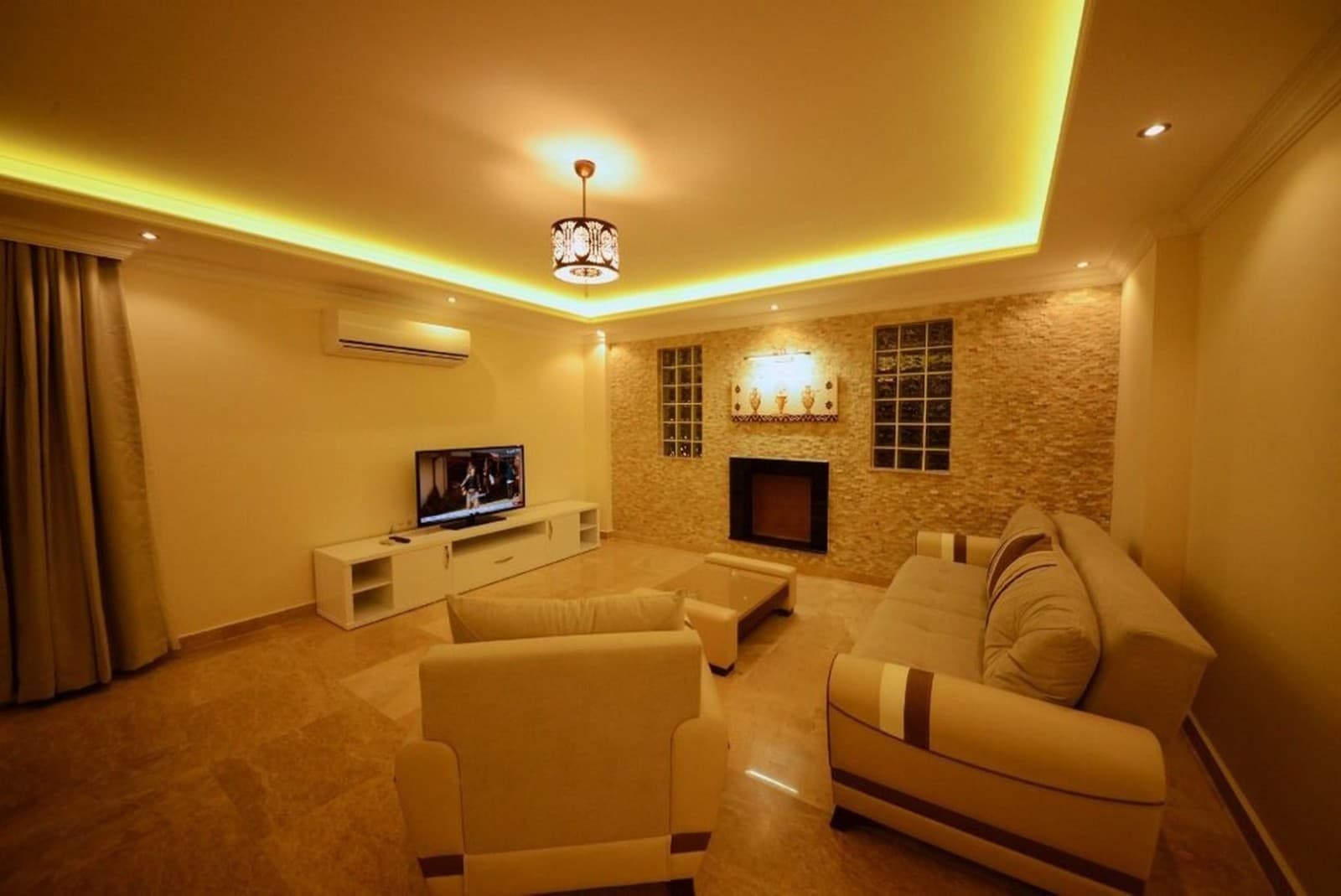 Bej koltuklar, TV ünitesi ve sarı tavan aydınlatmalı oturma odası.
