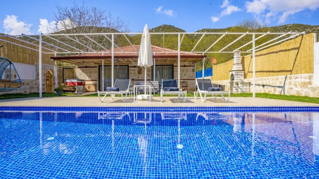 Mavi fayanslı yüzme havuzu, şezlonglar, beyaz güneş şemsiyesi ve arka planda taş villa ile dağ manzarası.