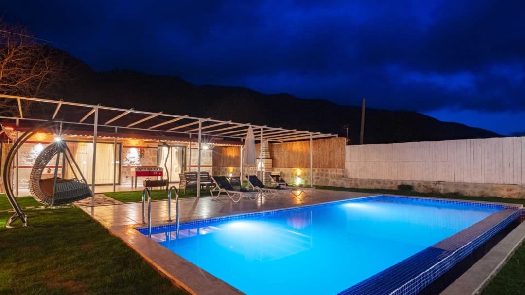 Işıklandırılmış havuz, şezlonglar, langırt ve bahçe salıncağının bulunduğu dağ manzaralı villa bahçesi.