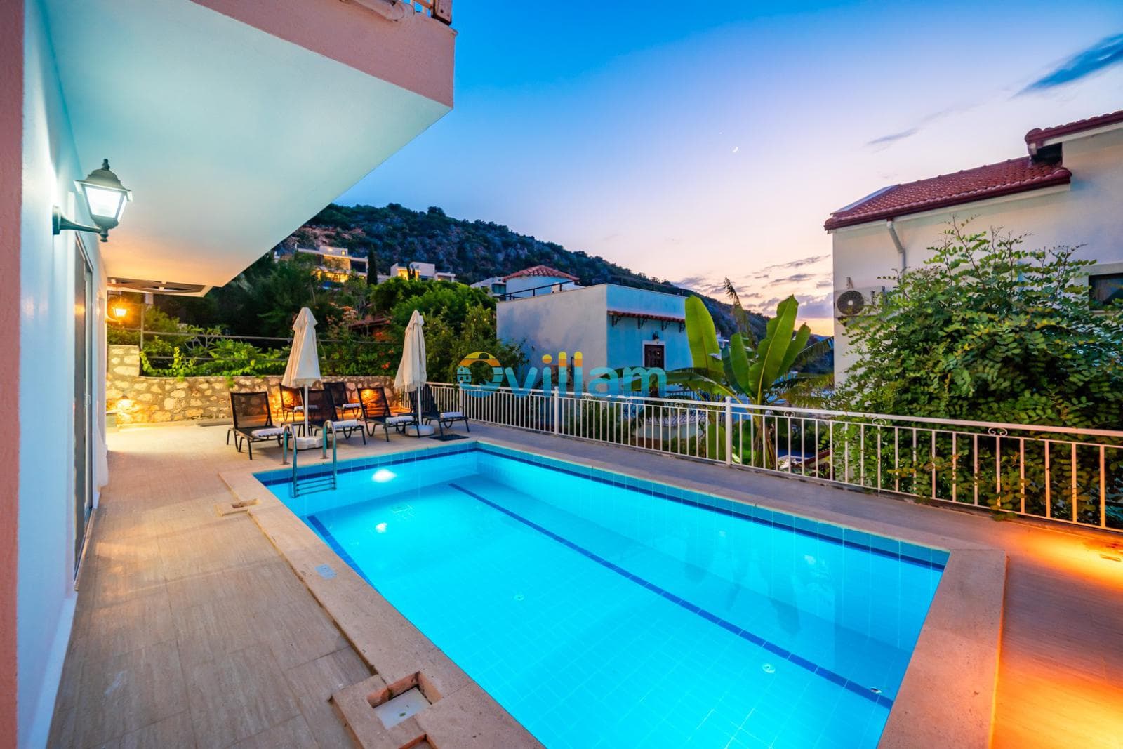 Akşam vakti aydınlatılmış havuz ve dağ manzaralı villa bahçesi.
