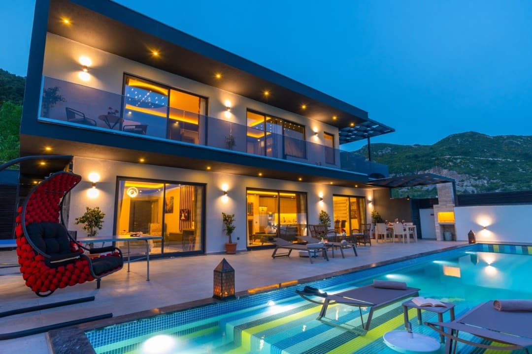Akşam ışıklandırmalı, havuzlu ve terasında şezlonglar ile kırmızı salıncak bulunan modern iki katlı villa.