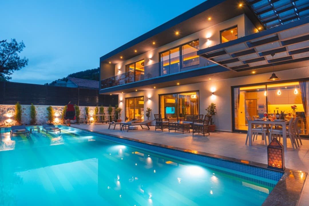 Işıklandırılmış havuz, şezlonglar ve yemek masası bulunan iki katlı modern villanın akşam görünümü.