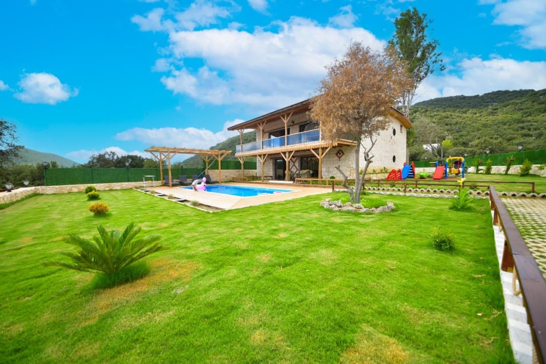 Geniş bahçeli, özel havuzlu, çocuk oyun parkı bulunan iki katlı taş villa ve dağ manzarası.
