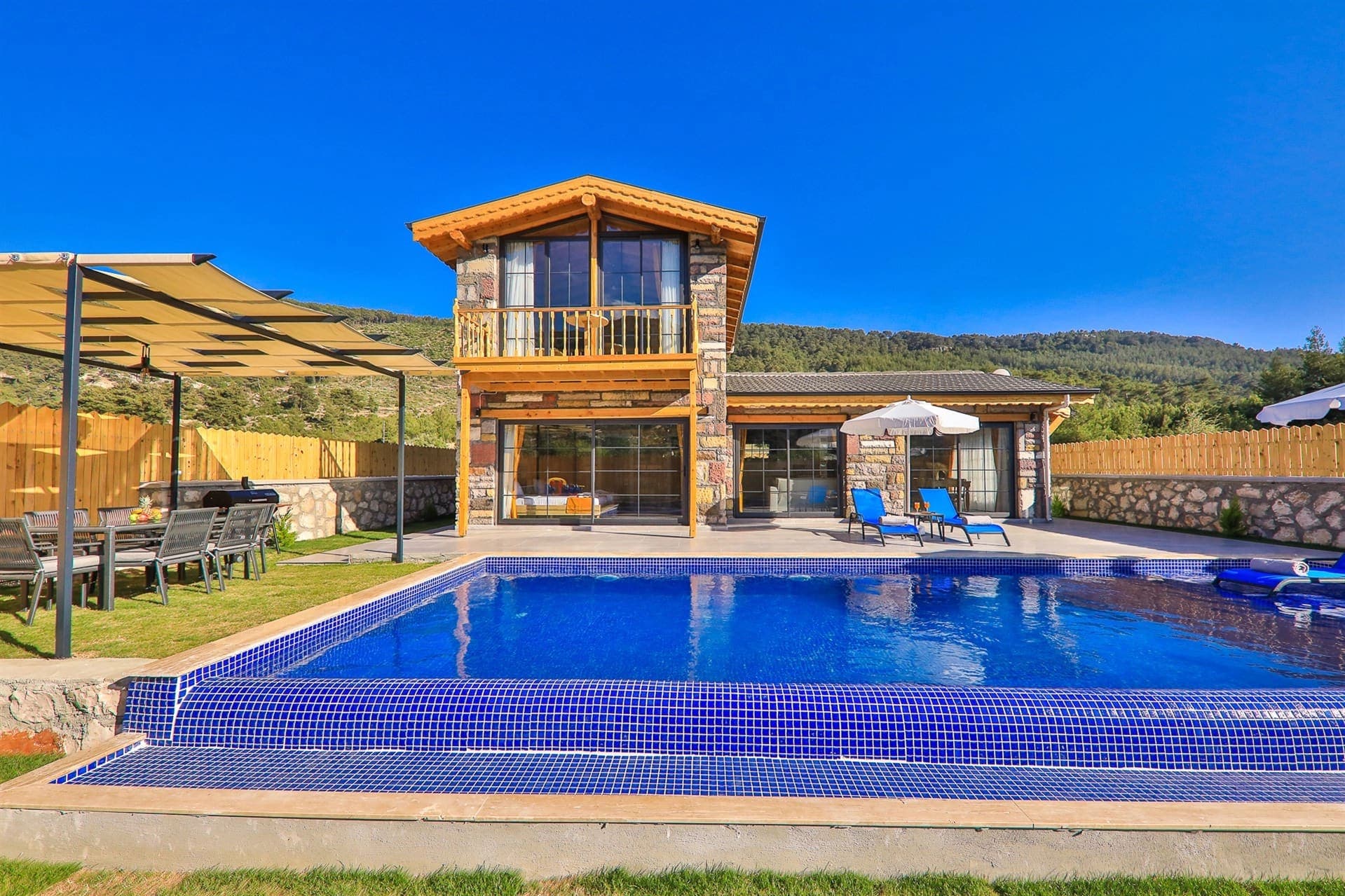 Önünde havuz, şezlonglar ve ahşap detaylı teras bulunan iki katlı taş villa.