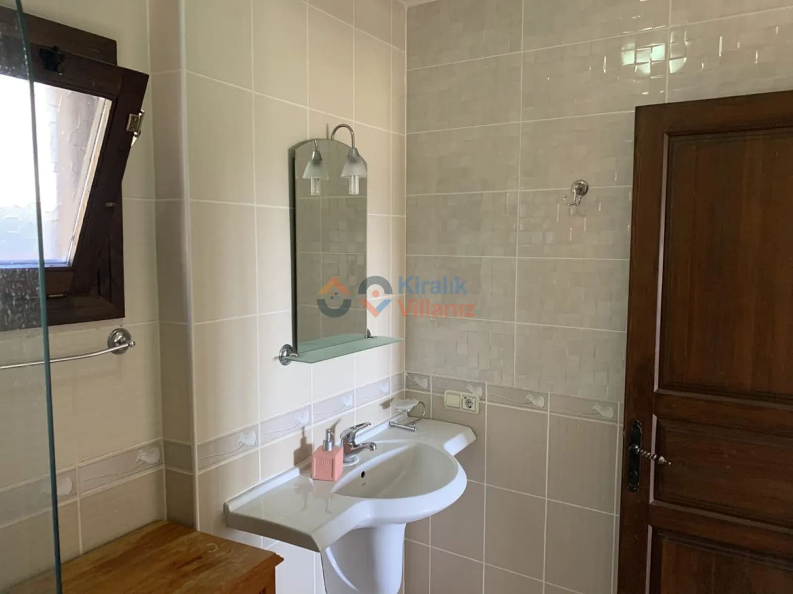 Bej seramik kaplı banyoda ayaklı lavabo, ayna ve ahşap pencere yer alıyor.