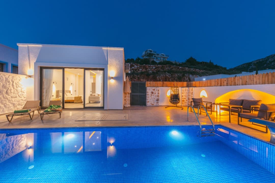 Gece ışıklandırmalı havuz kenarında şezlonglar ve oturma grubu bulunan tek katlı villa.