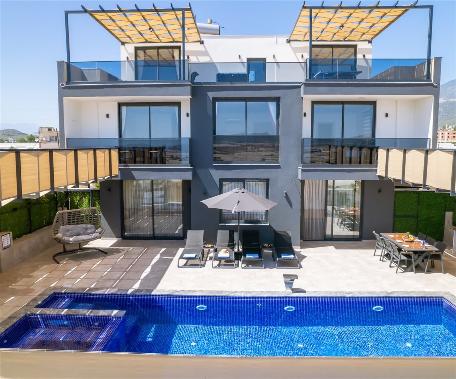 Güneşli günde, önünde havuz, şezlonglar ve yemek masası bulunan gri cepheli çok katlı villa.