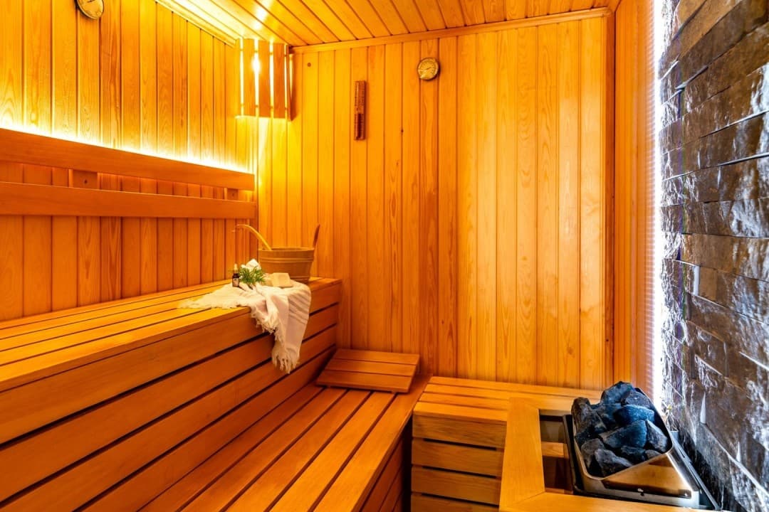 Ahşap kaplamalı, sauna sobası ve taş duvar detayları bulunan aydınlatmalı sauna odası.