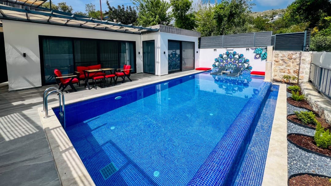 Mavi yüzme havuzu, verandada kırmızı minderli oturma grubu ve duvarda mandala deseni olan villa terası.