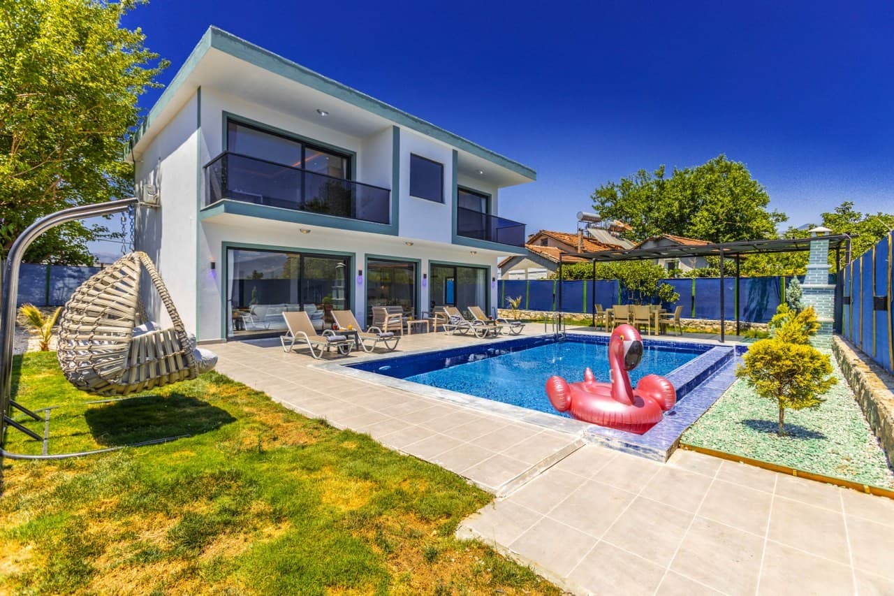 Önünde havuzu, şezlongları, salıncak koltuğu ve flamingo deniz yatağı bulunan iki katlı beyaz villa.