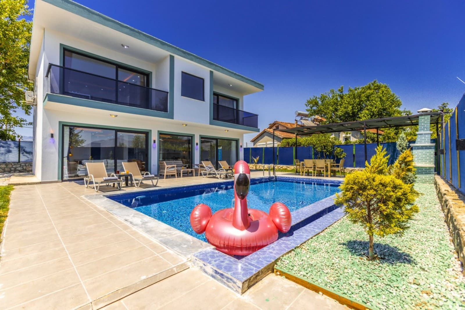 İki katlı beyaz villanın önünde mavi yüzme havuzu ve pembe flamingo deniz yatağı bulunuyor.
