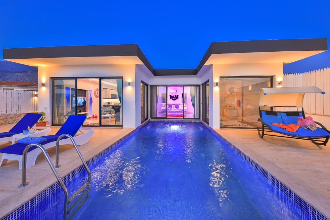 Aydınlatmalı yüzme havuzunun kenarında şezlonglar ve gün batımında modern villa.