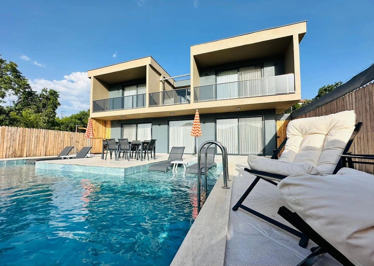 Açık havuz kenarında şezlonglar ve arka cephede iki katlı modern villa.