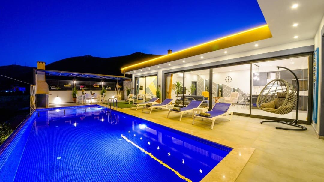 Gece aydınlatmalı havuz, şezlonglar, asma salıncak ve cam kapılı modern villanın dış görünümü.