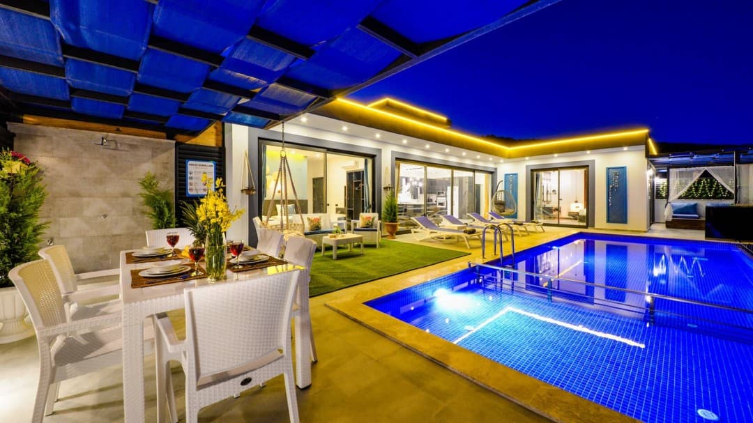 Mavi ışıklı yüzme havuzu, şezlonglar ve gölgelik altında yemek masası bulunan aydınlatılmış villa terası.