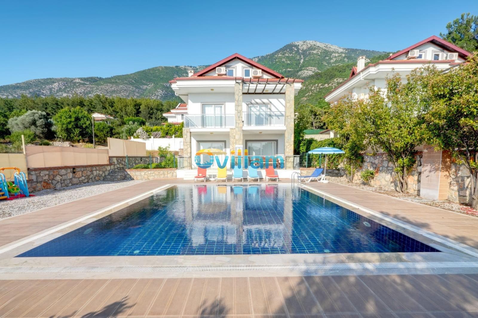 Dağ manzaralı geniş havuz ve önünde şezlonglar bulunan beyaz villa.