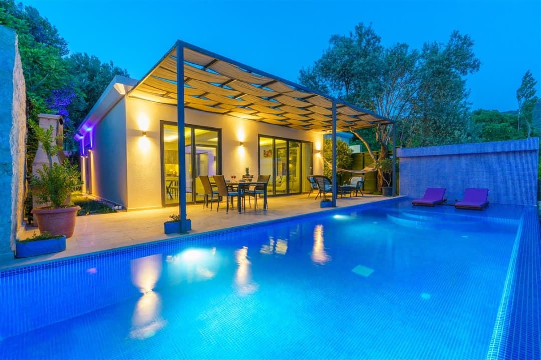 Akşam aydınlatmalı, havuz başında oturma grubu ve şezlong bulunan tek katlı villa.