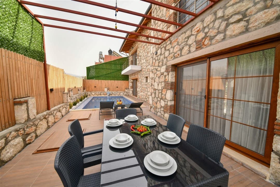 Taş duvarlı villanın terasındaki yemek masası seti, ahşap pergola ve arka planda özel yüzme havuzu.