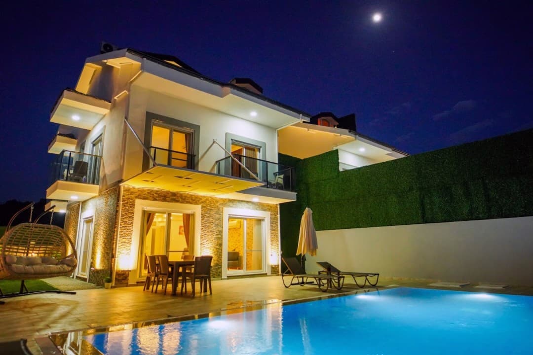 Gece çekiminde aydınlatılmış beyaz villanın havuzu ve bahçe oturma grubu.