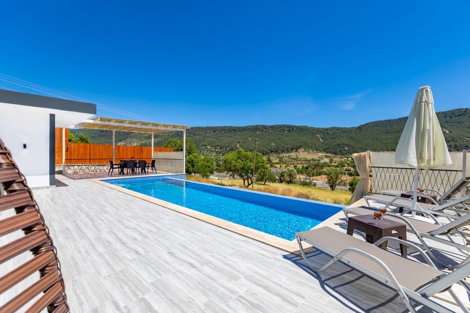 Güneşli bir günde, şezlonglar ve yemek masası bulunan, dağ manzaralı açık yüzme havuzlu villa terası.