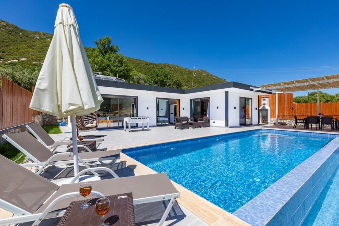 Mavi yüzme havuzu, şezlonglar ve arkasında yeşil dağ manzarası olan tek katlı modern villa.