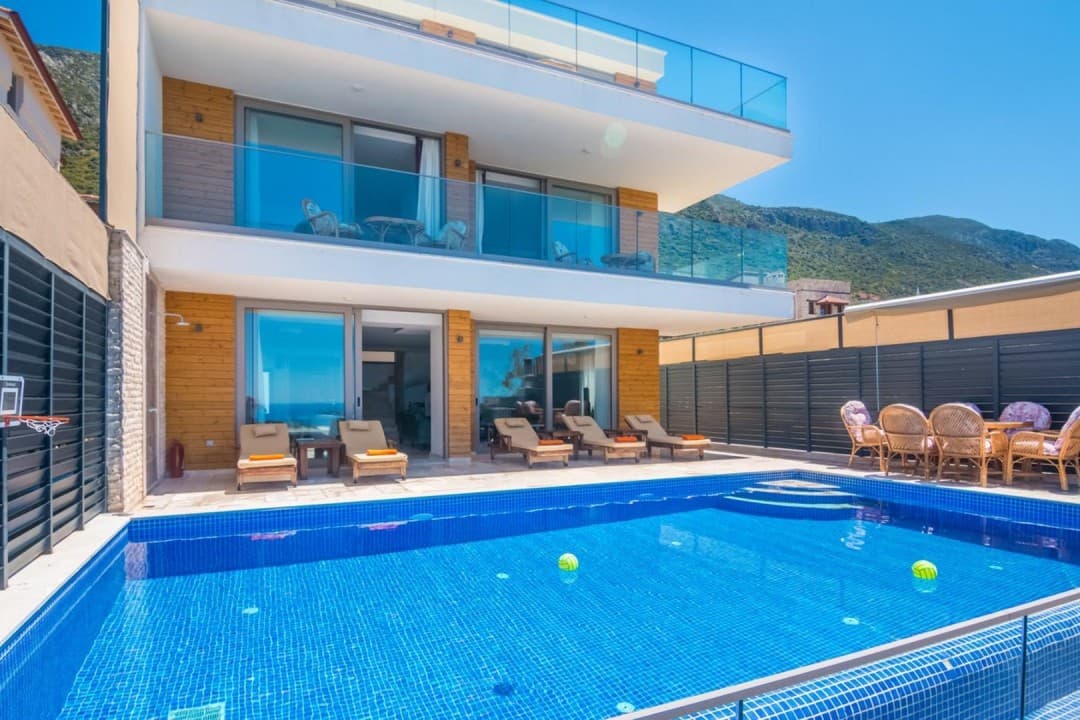 Mavi yüzme havuzu, şezlonglar ve masa grubu önünde yer alan cam balkonlu modern villa.