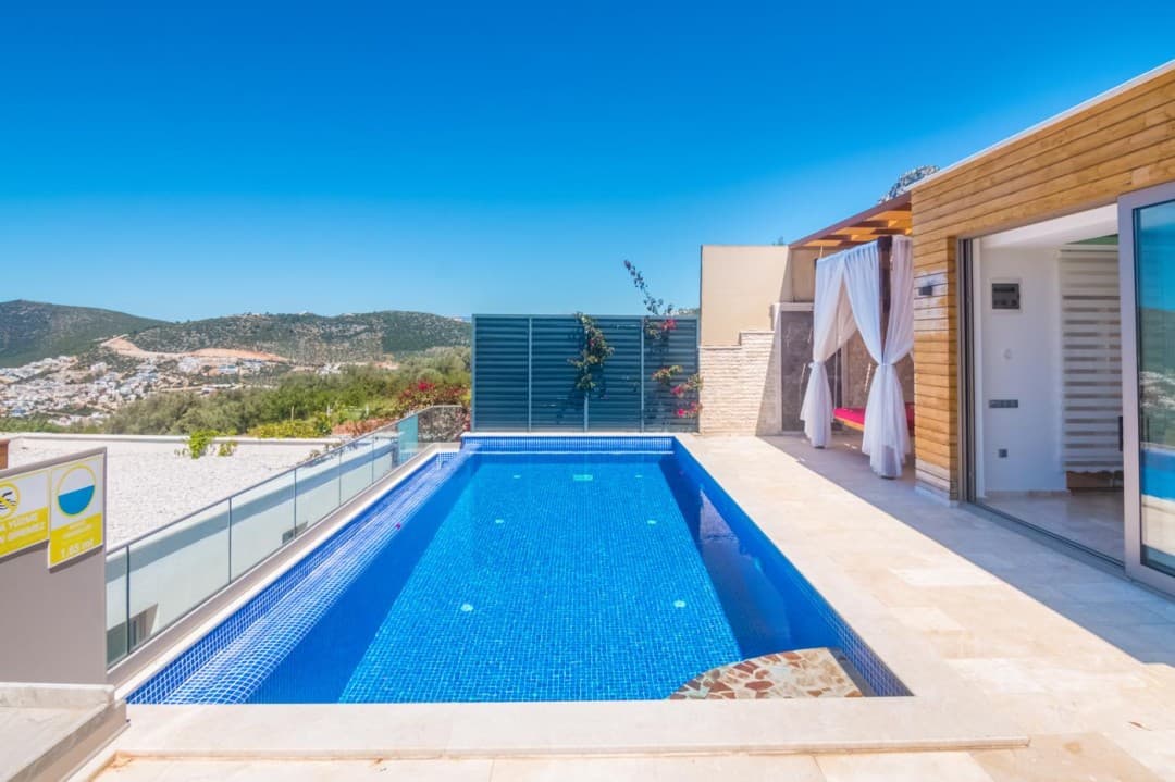 Dağ manzaralı terasta yer alan mavi yüzme havuzu, tül perdeli çardak ve ahşap kaplamalı villa.