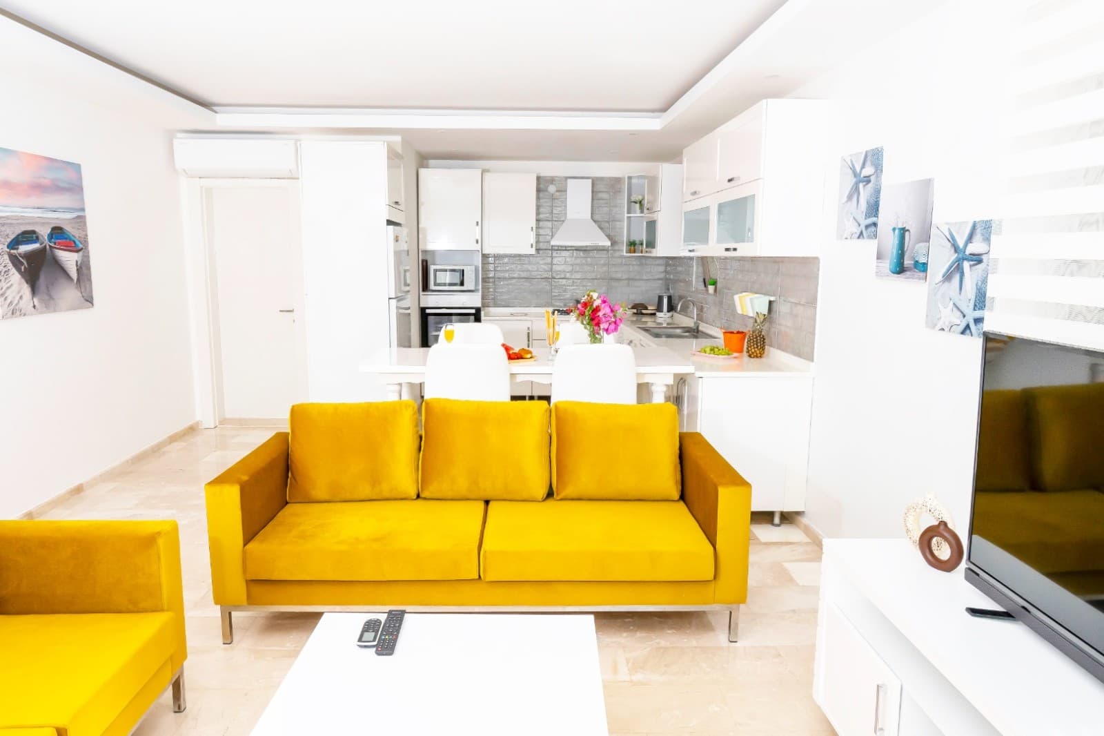 Açık renk zeminli, sarı koltuk takımı ve açık mutfağın bulunduğu salon.
