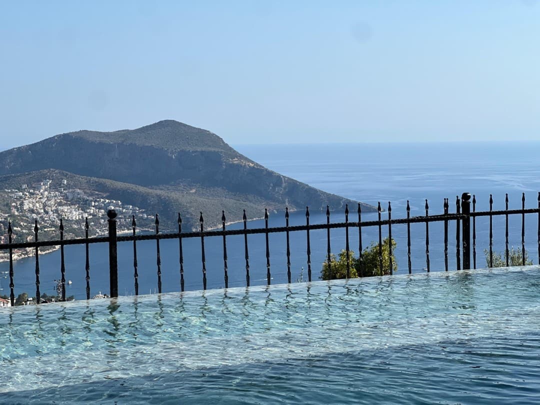 Havuz kenarından deniz, koy ve dağ manzarası sunan ferforje korkuluklu balkon.