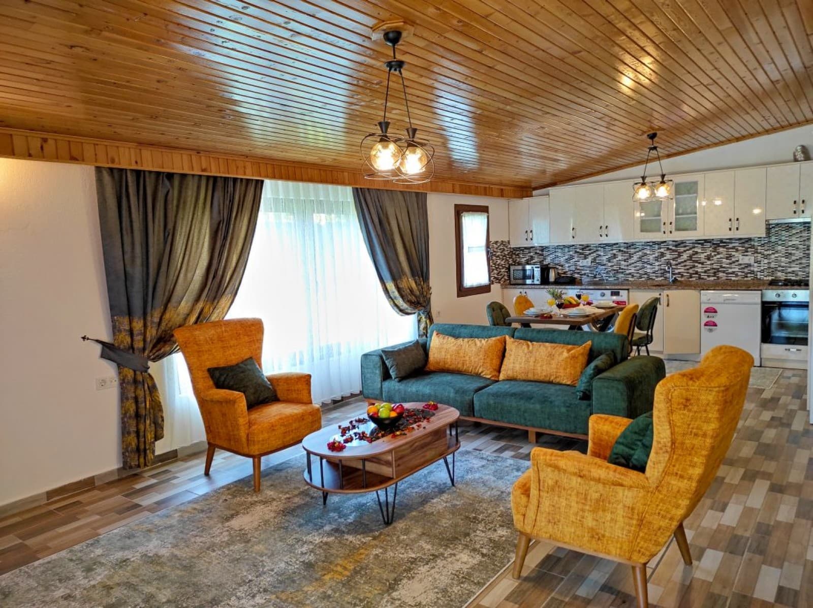 Ahşap tavanlı salonda yeşil kanepe, iki sarı berjer ve arka planda açık mutfak var.