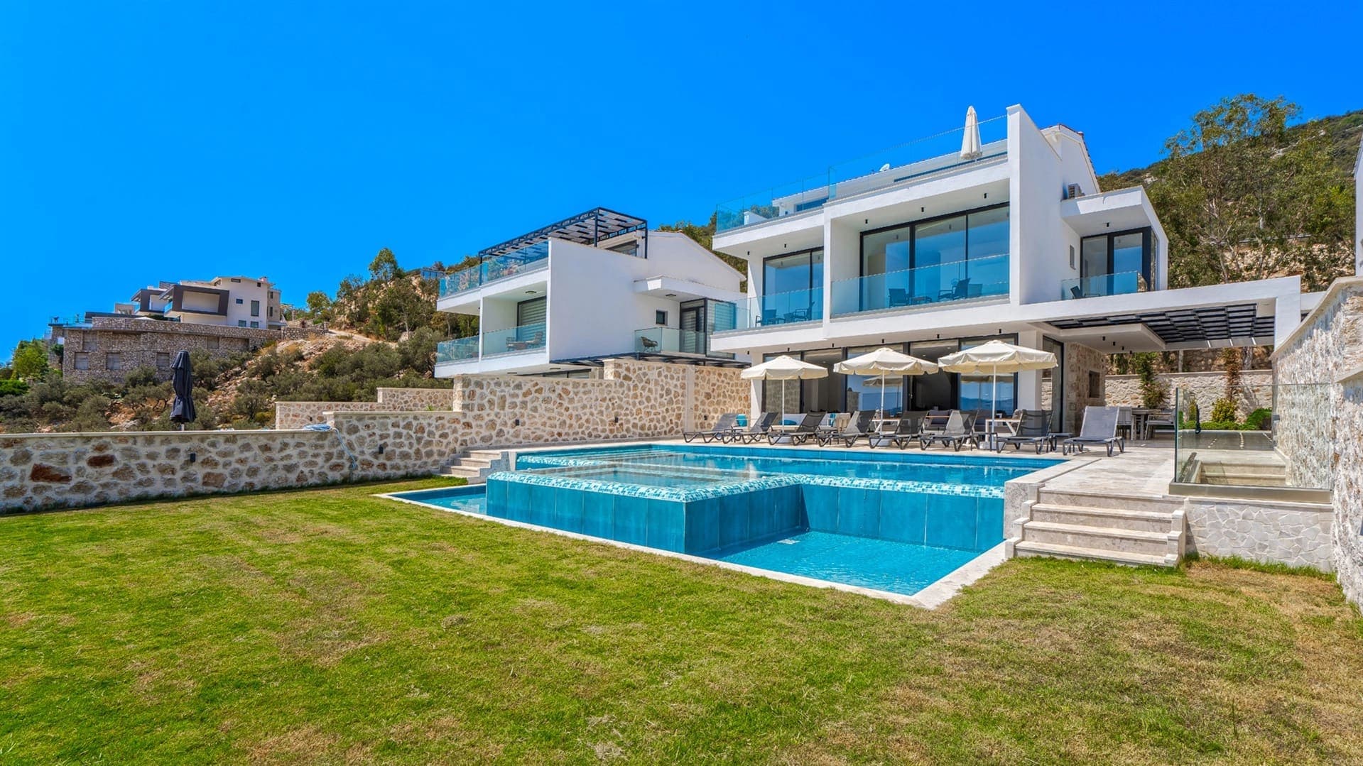 Çim alan ve şezlonglarla çevrili havuzun arkasında yer alan iki katlı beyaz villa.