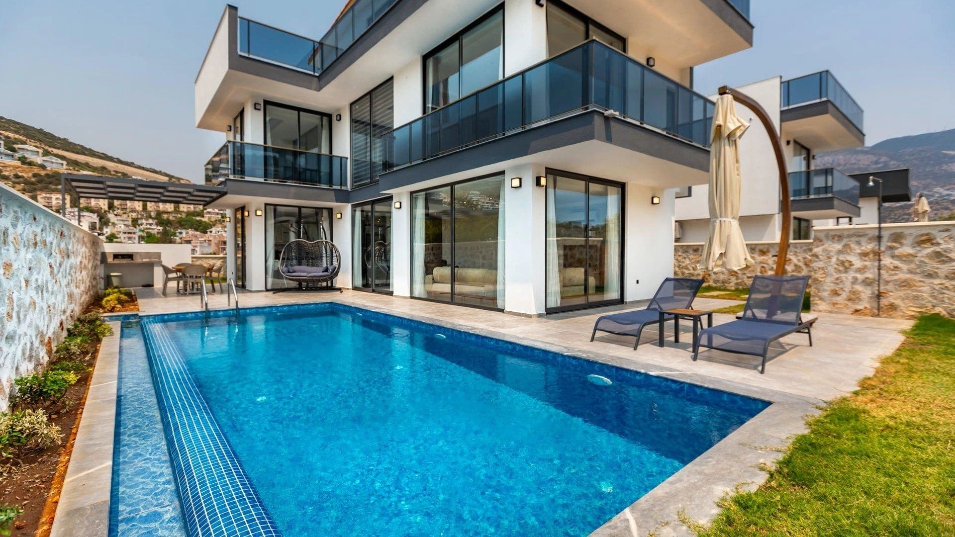 Modern villanın önünde yüzme havuzu, şezlonglar ve taş duvarlı bahçe var.