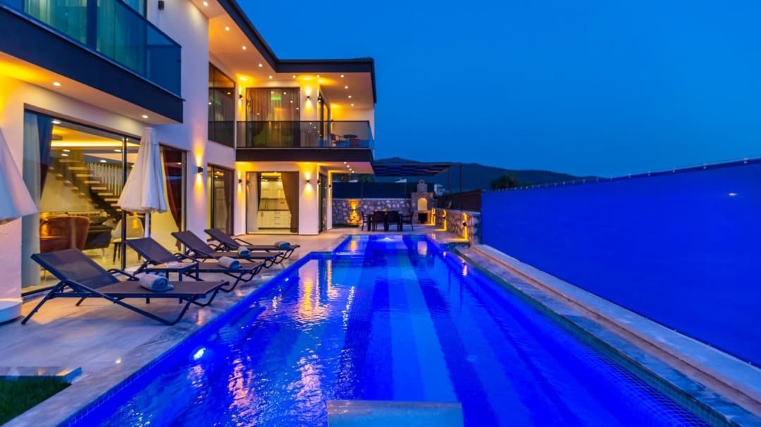 Akşam karanlığında dış cephesi aydınlatılmış iki katlı modern villanın önündeki uzun havuz ve şezlonglar.