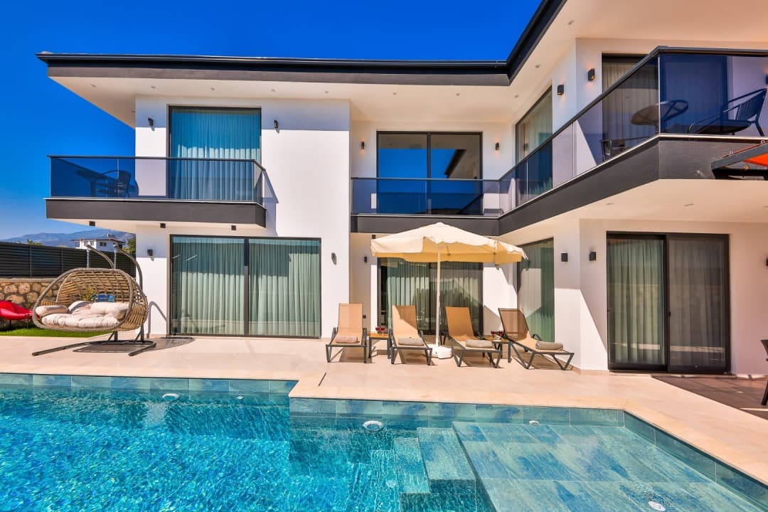 Gündüz vakti havuz kenarında şezlonglar, şemsiye ve rattan sallanan sandalye bulunan modern villa.