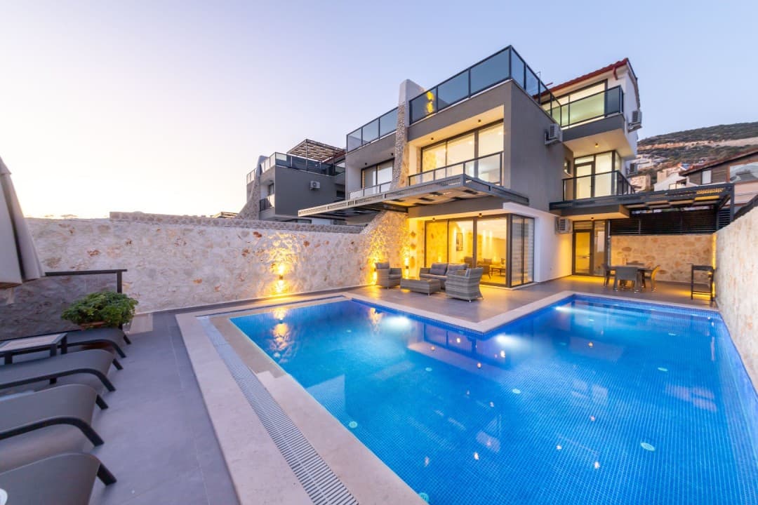 Akşam üstü ışıklandırılmış, havuzlu ve taş duvarlı modern iki katlı villa.