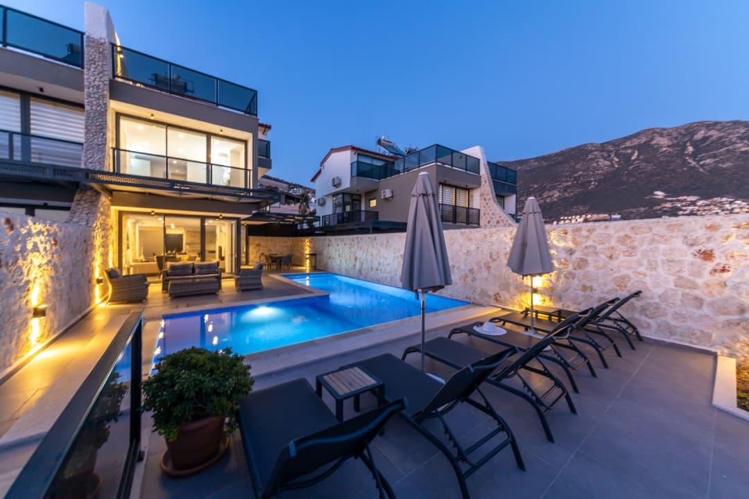Akşam vakti aydınlatmalı havuz kenarında şezlonglar ve arka planda dağ manzaralı villa.