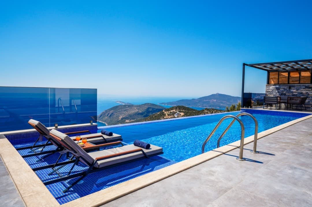 Masmavi deniz ve dağ manzarasına bakan sonsuzluk havuzu ve şezlonglar.