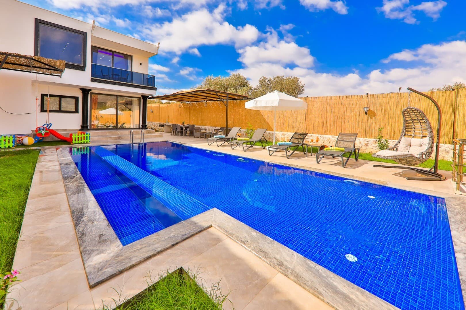 Mavi havuzlu, şezlonglu, salıncaklı ve çocuk oyun alanına sahip bahçeli beyaz modern villa.