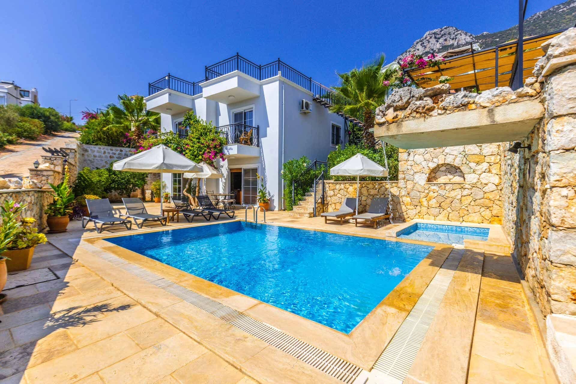 Dağ manzaralı taş duvarlı bahçe içinde havuz ve iki katlı beyaz villa.