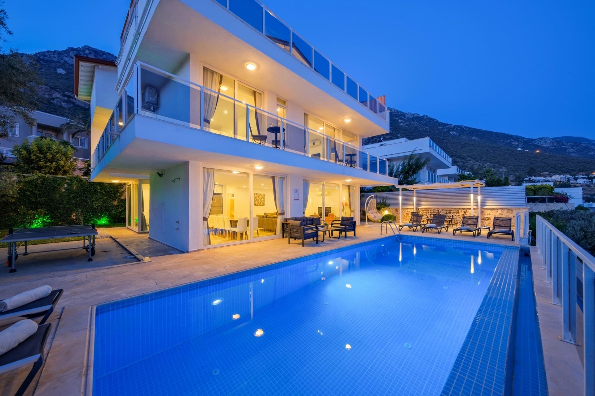 Aydınlatmalı havuz kenarında şezlonglar ve arka planda dağ manzaralı iki katlı villa.