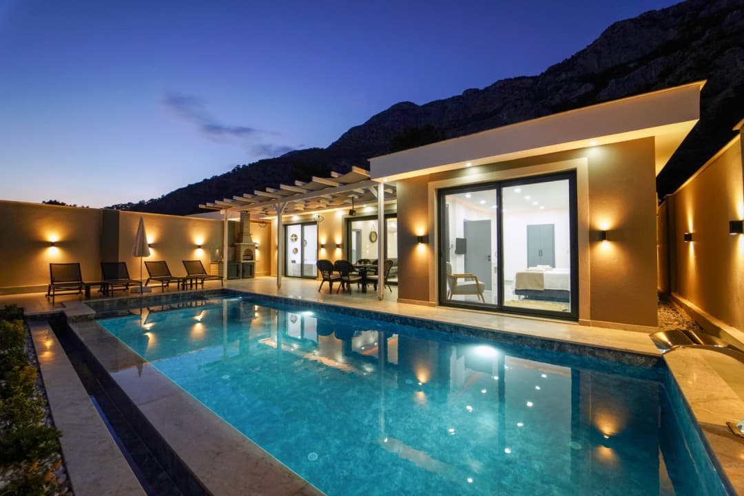 Akşam vakti havuz başı aydınlatmalı tek katlı villa ve arka planda dağ manzarası.