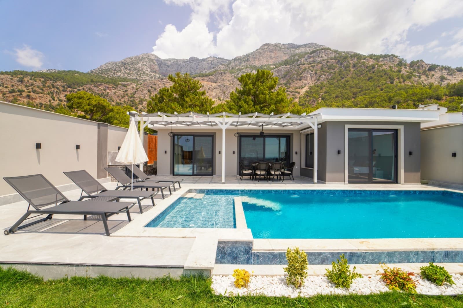 Dağ manzaralı, havuz kenarında şezlonglar ve gölgelikli oturma alanı bulunan villa dış cephesi.