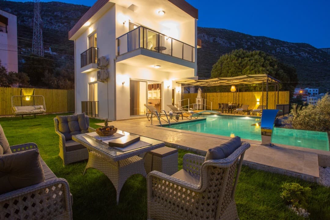Akşam ışıklandırmalı, bahçesinde yüzme havuzu ve oturma grubu bulunan iki katlı villa.